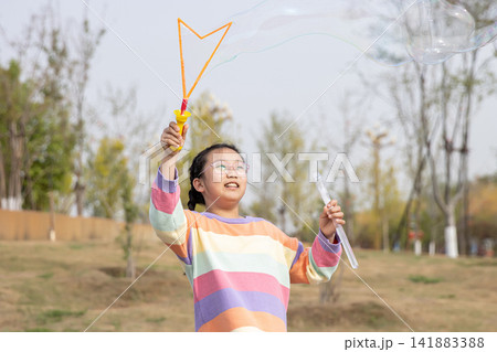Portrait of a smiling young girl in a colorful sweater playing with a large soap bubble outdoors. 141883388