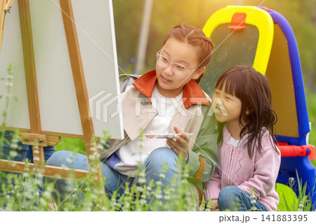 Two young Asian girls are sketching and painting at easels amidst the great outdoors, showcasing the beauty of both art education and nature. Two young Asian girls are sketching and painting at easels amidst the great outdoors, showcasing the beauty of both art education and nature. 141883395