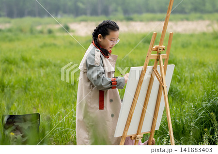 Young girl student painting on a canvas in a vast green meadow during art class. 141883403