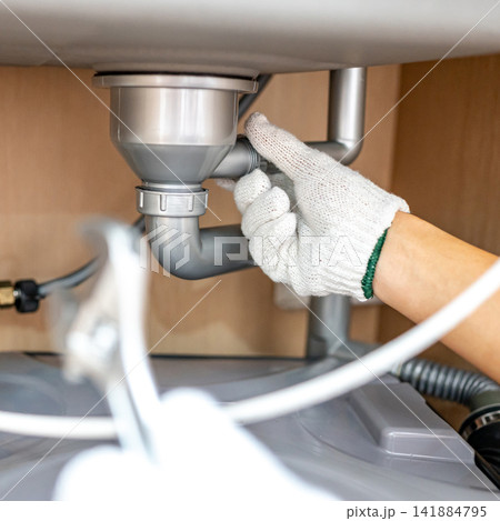 Plumber hand with glove fixing pipe connection under kitchen sink with adjustable wrench Plumber hand with glove fixing pipe connection under kitchen sink with adjustable wrench 141884795