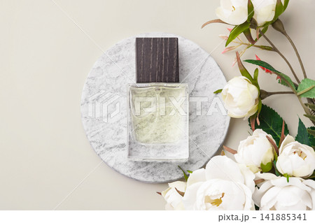 Perfume bottle on a round marble podium, decorated with decorative white roses and green leaves on beige background Perfume bottle on a round marble podium, decorated with decorative white roses and green leaves on beige background 141885341