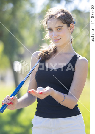 Young woman holding golf ball while standing on grass with golf club in hand during sunny day in park Young woman holding golf ball while standing on grass with golf club in hand during sunny day in park 141885482