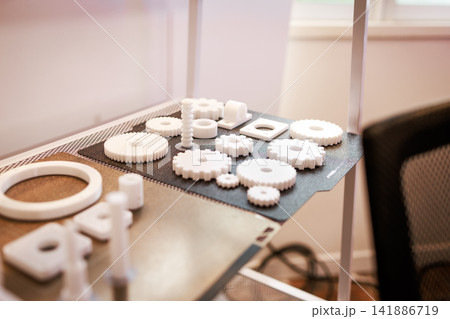 Close-up of white 3d printed gears and small parts arranged on a workbench in a workshop, with soft natural light and a blurred background Close-up of white 3d printed gears and small parts arranged on a workbench in a workshop, with soft natural light and a blurred background 141886719
