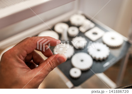 Close up of a hand holding a small white gear with other gears blurred in the background, repair, mechanics and precision work. Close up of a hand holding a small white gear with other gears blurred in the background, repair, mechanics and precision work. 141886720