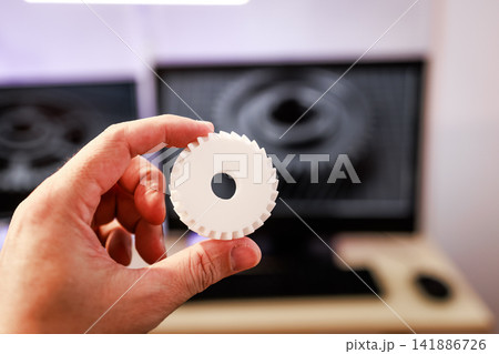Hand holding a white 3d printed gear in front of a blurred computer screen in a workshop or design office 141886726