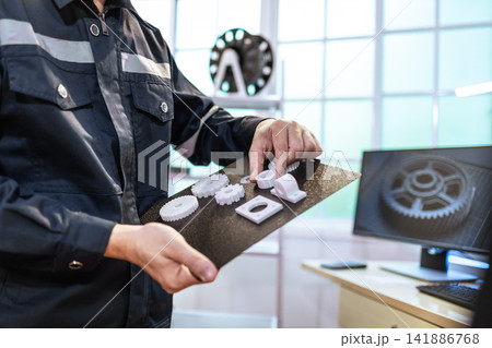 Technician holding a tray with 3d printed parts in a workshop, with a computer screen a gear model in the background Technician holding a tray with 3d printed parts in a workshop, with a computer screen a gear model in the background 141886768