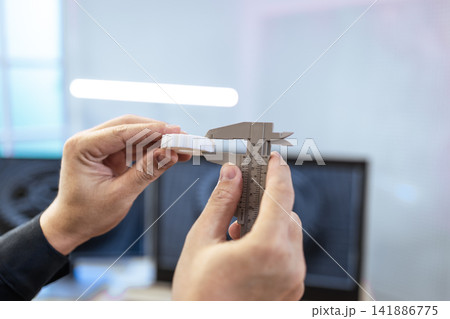 Hands measuring a small white part with a caliper in an office setting, precision, inspection, and quality control Hands measuring a small white part with a caliper in an office setting, precision, inspection, and quality control 141886775
