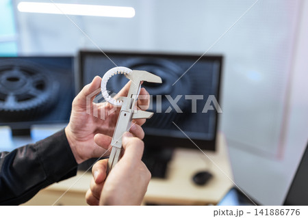 Hands measuring a 3d printed gear with caliper in a workshop, with blurred computer screens in the background Hands measuring a 3d printed gear with caliper in a workshop, with blurred computer screens in the background 141886776