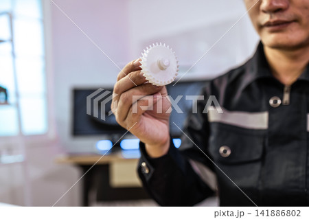 Man in work wear holding a white plastic gear in a workshop, a machine part for repair or manufacturing. Man in work wear holding a white plastic gear in a workshop, a machine part for repair or manufacturing. 141886802