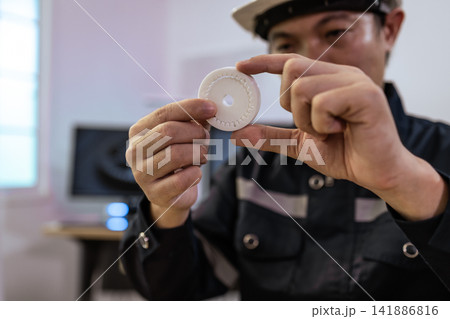 Worker in uniform inspecting a small white plastic part with hands in a workshop close up 141886816