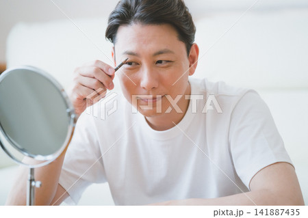 卓上鏡を見ながらアイブロウペンシルで眉毛を描く微笑みのミドル男性のメンズメイクアップ 141887435