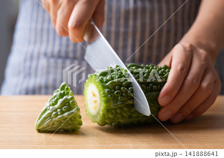 Woman hands holding knife and cutting fresh bitter gourd on wooden board, preparing food ingredient Woman hands holding knife and cutting fresh bitter gourd on wooden board, preparing food ingredient 141888641