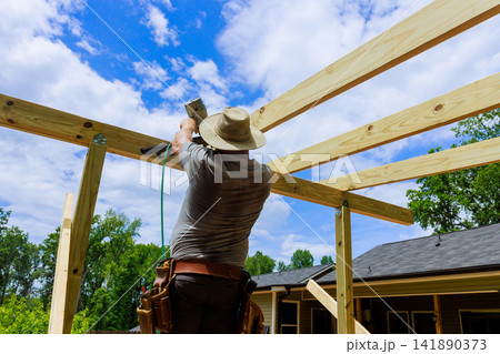 Carpenter uses pneumatic nail gun to attach wooden beams in construction project at house 141890373