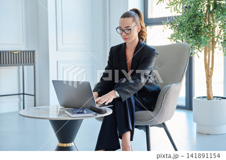 Young focused businesswoman working on laptop at small table in modern office Young focused businesswoman working on laptop at small table in modern office 141891154