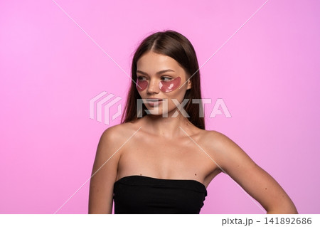 Young woman applying cosmetic eye patches for under eye skin care, looking away on pink studio background Young woman applying cosmetic eye patches for under eye skin care, looking away on pink studio background 141892686