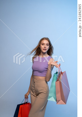 Young woman carrying colorful shopping bags on a light blue background, representing consumerism, fashion, and discount deals Young woman carrying colorful shopping bags on a light blue background, representing consumerism, fashion, and discount deals 141892905