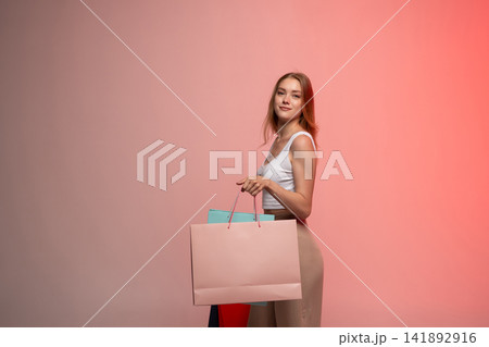 Young woman smiling, holding multiple shopping bags, enjoying retail therapy and consumerism against a pink background Young woman smiling, holding multiple shopping bags, enjoying retail therapy and consumerism against a pink background 141892916
