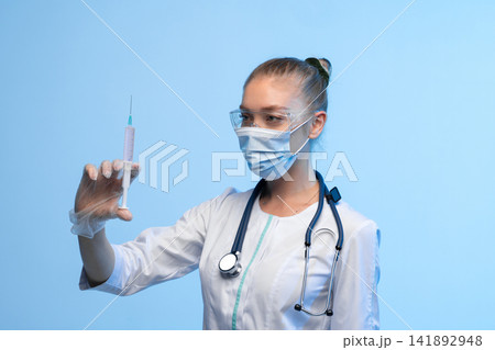 Healthcare professional wearing protective mask, safety goggles, and medical uniform, holding a syringe filled with pink liquid Healthcare professional wearing protective mask, safety goggles, and medical uniform, holding a syringe filled with pink liquid 141892948