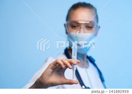 Medical professional wearing mask and safety glasses holding a syringe with vaccine for injection or treatment 141892959
