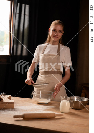 Young woman sifting flour into a bowl, preparing ingredients for baking in her kitchen, making homemade dough Young woman sifting flour into a bowl, preparing ingredients for baking in her kitchen, making homemade dough 141893332