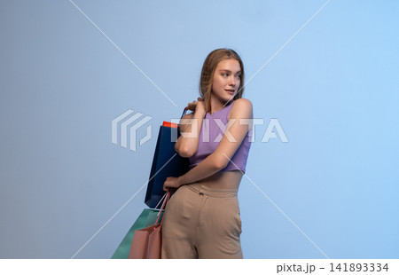 Young woman enjoying shopping, carrying colorful bags, looking away and smiling on a blue background Young woman enjoying shopping, carrying colorful bags, looking away and smiling on a blue background 141893334