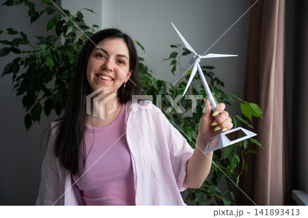 Smiling woman holding a small model wind turbine in a room with indoor plants. Green energy advocacy and eco-friendly lifestyle awareness Smiling woman holding a small model wind turbine in a room with indoor plants. Green energy advocacy and eco-friendly lifestyle awareness 141893413