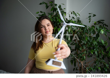 Young woman presenting a miniature wind turbine against a green indoor backdrop. Renewable energy innovation and eco-friendly startup campaign 141893426