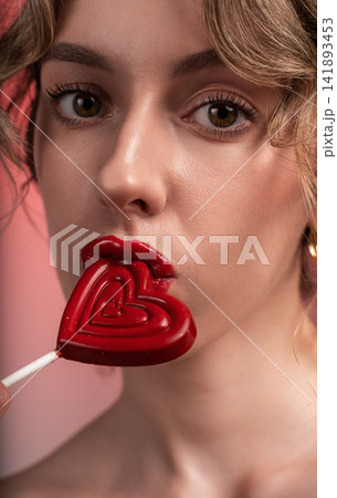 Woman holding red heart lollipop with glossy lips and looking at camera, celebrating sweet love and romance Woman holding red heart lollipop with glossy lips and looking at camera, celebrating sweet love and romance 141893453