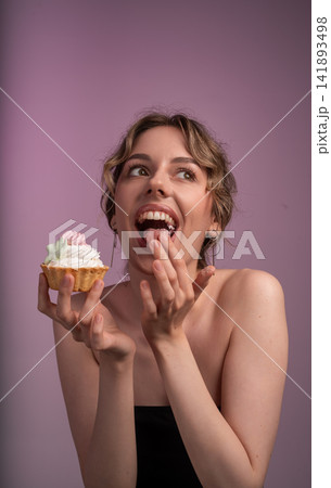 Woman enjoying a cupcake, licking finger with sweet cream, feeling temptation and pleasure, studio background Woman enjoying a cupcake, licking finger with sweet cream, feeling temptation and pleasure, studio background 141893498