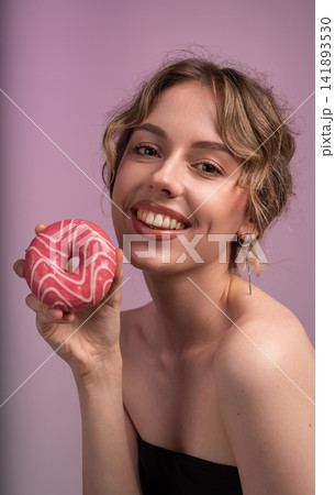 Young woman smiling and holding a delicious pink glazed donut, symbolizing joy, indulgence, and sweet treat pleasure Young woman smiling and holding a delicious pink glazed donut, symbolizing joy, indulgence, and sweet treat pleasure 141893530