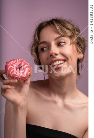 Happy young woman holding glazed pink donut and smiling, enjoying sweet dessert against purple background Happy young woman holding glazed pink donut and smiling, enjoying sweet dessert against purple background 141893531