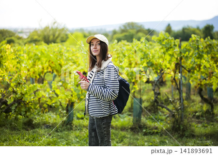 Young woman standing in a vineyard and holding a smartphone, illustrating the depth and perspective of seasonal vineyard rows. Young woman standing in a vineyard and holding a smartphone, illustrating the depth and perspective of seasonal vineyard rows. 141893691