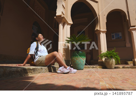 Woman relaxing in sun at historic Tangier courtyard with backpack and potted plants 141893787