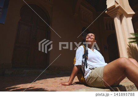 Smiling woman traveler sitting in the sun by a historic Tangier Morocco building with backpack 141893788