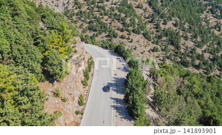 Aerial view of black car driving along winding asphalt mountain road through green pine forest in Lefka Ori region on Crete island under sunny day Greece 141893901