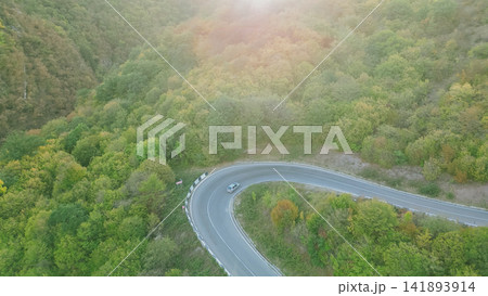 High angle aerial view of single silver car driving on sharp hairpin curve road through mountain forest illuminated by soft sunset light in Georgia High angle aerial view of single silver car driving on sharp hairpin curve road through mountain forest illuminated by soft sunset light in Georgia 141893914