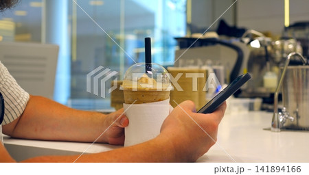 Hands holding cold drink and smartphone at cafe counter, clear cup with dome lid and black straw, iced beverage wrapped in napkin beside blurred espresso machines and bright interior Hands holding cold drink and smartphone at cafe counter, clear cup with dome lid and black straw, iced beverage wrapped in napkin beside blurred espresso machines and bright interior 141894166