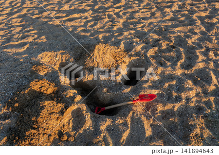 Red toy shovel in dug sand hole on beach at golden sunset Red toy shovel in dug sand hole on beach at golden sunset 141894643