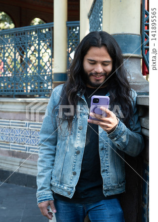 Young man smiling using smartphone outdoor with long hair Young man smiling using smartphone outdoor with long hair 141895565
