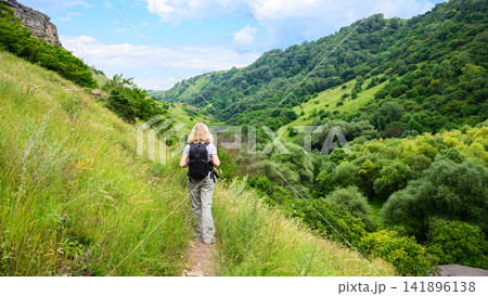 Blonde woman hiking on a mountain trail with a backpack. Female hiker walking along a grassy slope overlooking a lush green valley. Outdoor adventure and active lifestyle concept Blonde woman hiking on a mountain trail with a backpack. Female hiker walking along a grassy slope overlooking a lush green valley. Outdoor adventure and active lifestyle concept 141896138