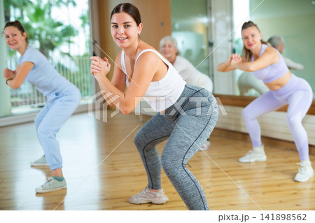 Four emotional fit young mature women in sportswear warming up with squats before aerobic dance training in gym Four emotional fit young mature women in sportswear warming up with squats before aerobic dance training in gym 141898562