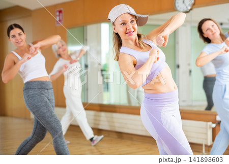 Joyful young mature women in sportswear learning new movements with hands at chest level while dancing together in hall Joyful young mature women in sportswear learning new movements with hands at chest level while dancing together in hall 141898603