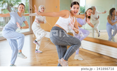 Class of happy women of different ages in sportswear training with knee up while learning dance moves with trainer in gym Class of happy women of different ages in sportswear training with knee up while learning dance moves with trainer in gym 141898718