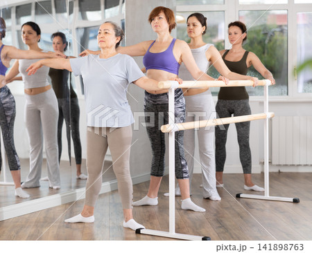 Women of different ages exercise near the ballet barre at group training session in batman batyu position Women of different ages exercise near the ballet barre at group training session in batman batyu position 141898763