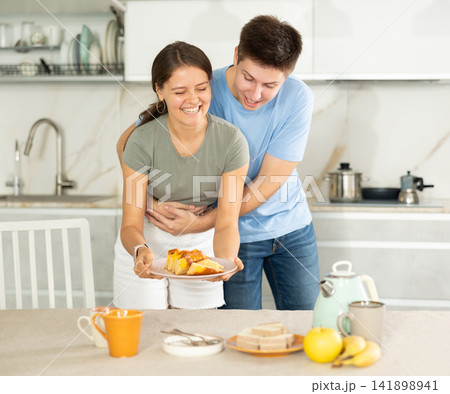 Guy gently hugs girlfriend around waist during preparations for afternoon snack Guy gently hugs girlfriend around waist during preparations for afternoon snack 141898941