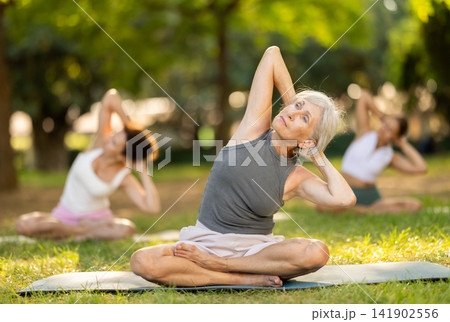 Happy old lady and class of calm sports women in activewear stretching muscles and doing tilts while sitting in lotus pose on mats during yoga training together at city park 141902556