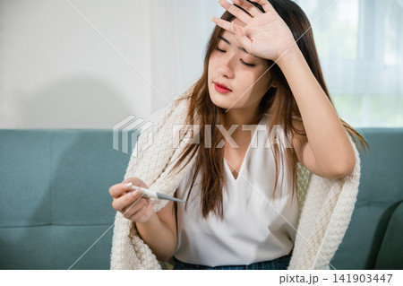 Sick Asian young woman sit under blanket on sofa she sneeze with digital thermometer Sick Asian young woman sit under blanket on sofa she sneeze with digital thermometer 141903447
