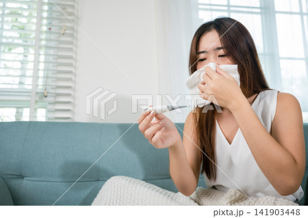 Sick Asian young woman sit under blanket on sofa she sneeze with digital thermometer Sick Asian young woman sit under blanket on sofa she sneeze with digital thermometer 141903448