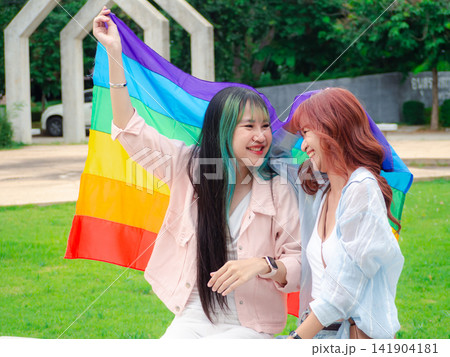Happy Asian young LGBTQ lesbian couple laughing with rainbow pride flag at park. Gen Z female lovers smiling, enjoying romantic relationships, freedom, gender diversity, equality. Happy Asian young LGBTQ lesbian couple laughing with rainbow pride flag at park. Gen Z female lovers smiling, enjoying romantic relationships, freedom, gender diversity, equality. 141904181