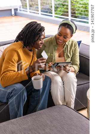 African American couple sitting on balcony, man in mustard sweater holding card, woman using tablet 141907707